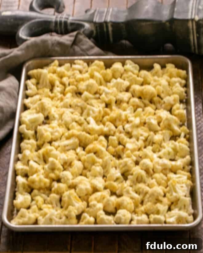 Cauliflower florets spread out in a single, uncrowded layer on a large baking sheet, seasoned and ready for roasting.