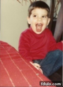 The birthday boy, Tom, as a child in a red shirt and jeans with a big smile, ready to eat his cake.