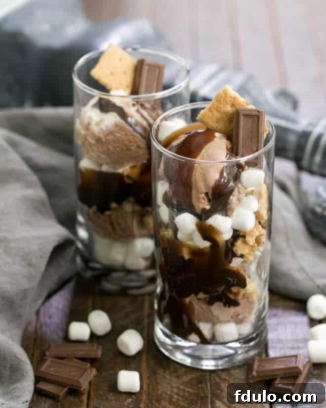 A vibrant S'mores Ice Cream Sundae presented in a clear glass, with visible layers of creamy ice cream, golden marshmallows, crunchy graham crackers, and rich fudge, garnished with chocolate squares and extra marshmallows.
