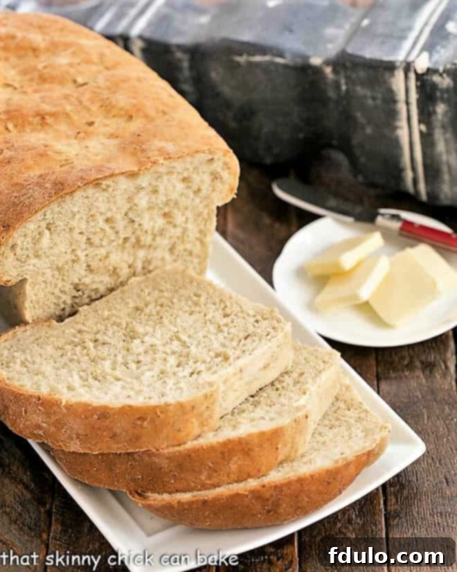 Oat Bread Recipe - a sweet, hearty bread flavored with a touch of brown sugar A partially sliced loaf of wholesome oatmeal bread rests on a white serving tray, with a small plate of butter pats softly blurred in the background, inviting guests to enjoy.