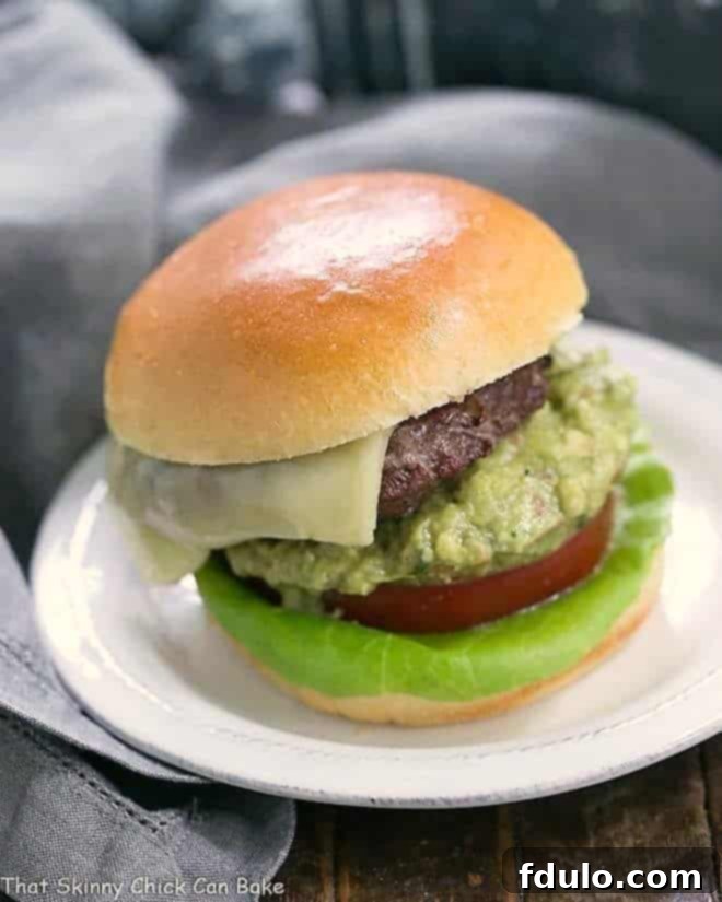 Grilled Guacamole Burger - a grilled beef hamburger topped with melted Monterey jack cheese and a creamy mound of guacamole A delicious Grilled Guacamole Burger, also known as an Avocado Burger, served on a white ceramic plate. It's topped with melted Monterey Jack cheese, a generous scoop of fresh guacamole, vibrant red tomato, and crisp green lettuce.