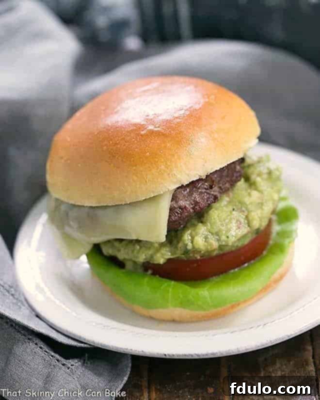 Guac Grilled Burgers 2 A beautifully presented Grilled Guacamole Burger on a round white plate, garnished with fresh lettuce, a thick slice of tomato, and a generous dollop of creamy guacamole over melted cheese.
