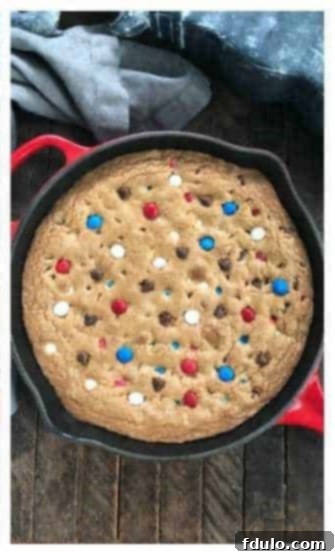 A warm skillet cookie with a hand pressing extra red, white, and blue M&M's and chocolate chips onto its surface.