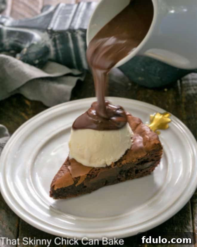 Slice of brownie tart topped with a scoop of vanilla ice cream with chocolate sauce pouring out from a white ceramic pitcher.