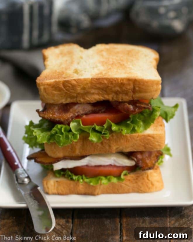 A glorious double-decker Brie Bacon Lettuce and Tomato Sandwich, perfectly layered on a square white plate with a small red handled knife.