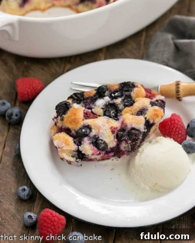 A serving of Easy Mixed Berry Cobbler on a white dessert plate with a scoop of ice cream and fresh berries, showcasing its golden crust and vibrant fruit filling.