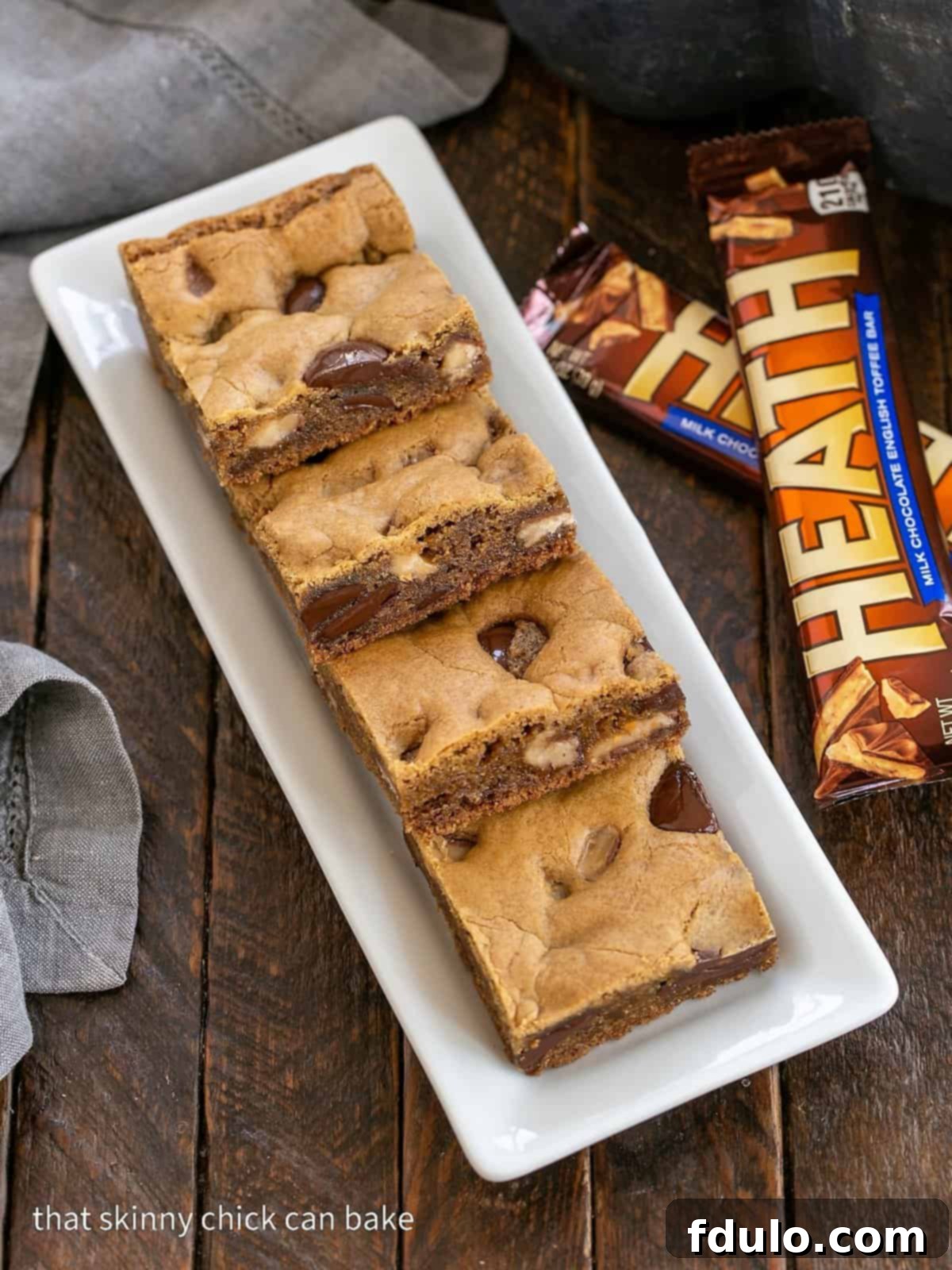 An inviting overhead view of freshly baked Chocolate Chunk Blondies, generously studded with glistening toffee chunks, presented on a rustic white ceramic tray. The golden-brown edges and soft center promise a delectable experience.