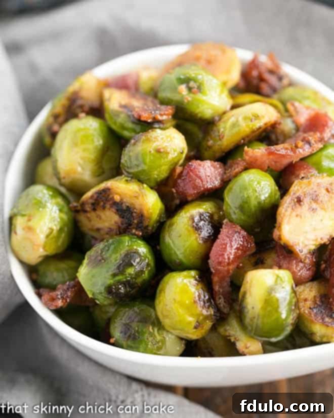 Closeup of perfectly charred Maple Syrup Mustard Glazed Brussels Sprouts glistening with glaze and dotted with crispy bacon in a white ceramic bowl.
