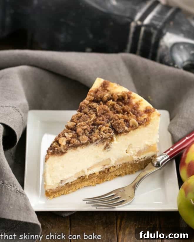 Slice of Apple Crisp Cheesecake on a white plate with a red handled fork, showcasing layers of graham cracker crust, spiced apples, creamy cheesecake, and oat streusel topping.