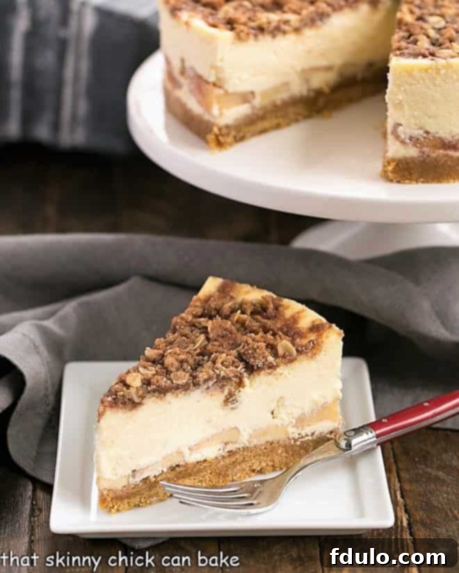 Slice of Apple Crisp Cheesecake presented beautifully in front of a full cheesecake on a cake stand.