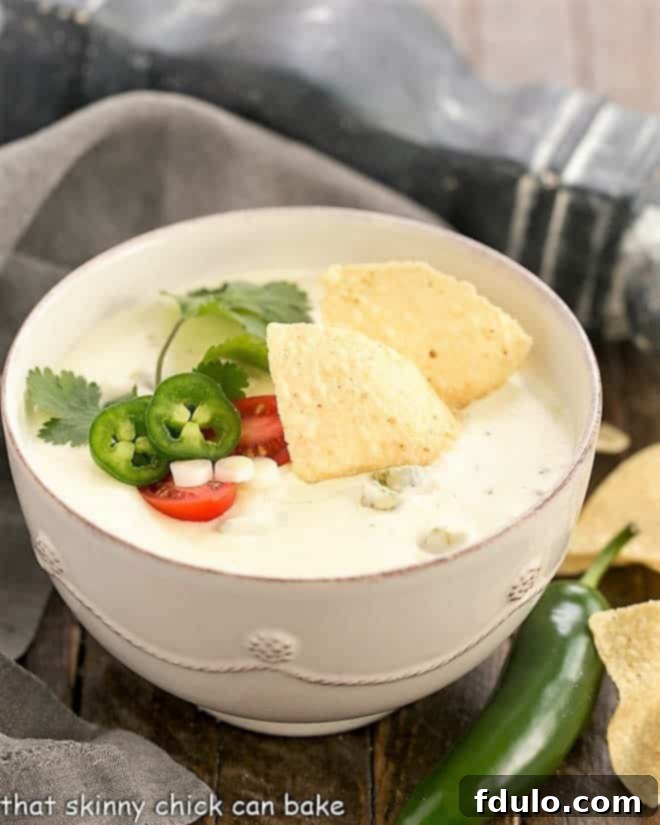 Easy Homemade Queso Dip in a white serving bowl topped with tortilla chips, jalapeno slices, cilantro sprig, showcasing a rich and creamy texture.