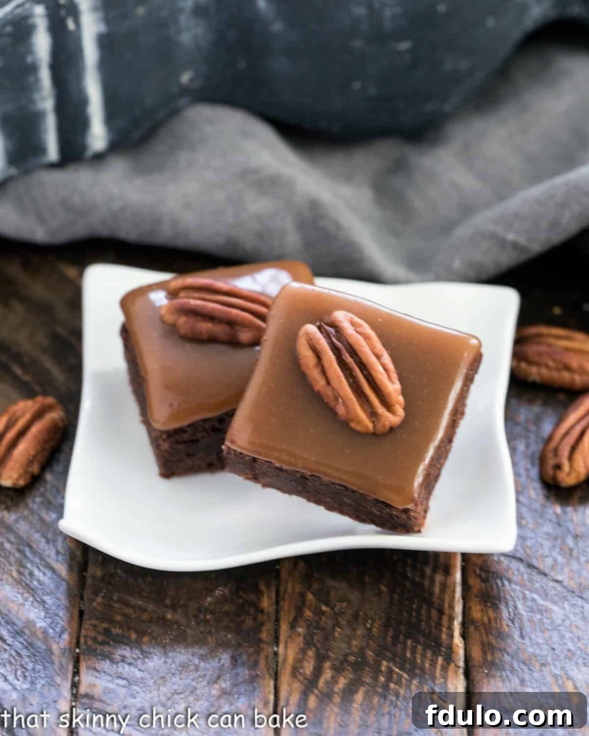 Two glistening Ultimate Turtle Brownies, each topped with golden caramel and a pecan half, resting on a small white wavy plate, showcasing their irresistible layers and tempting texture.