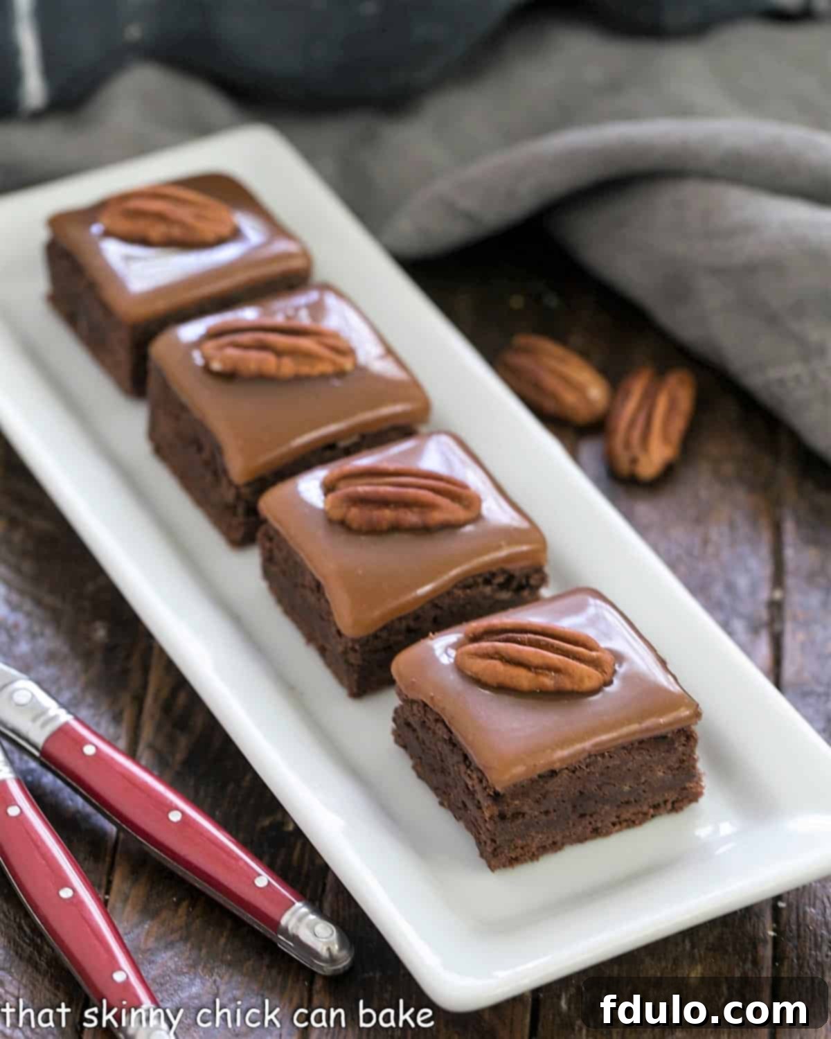 A white rectangular tray filled with freshly baked Ultimate Turtle Brownies, glistening with perfectly set caramel and adorned with crunchy pecan halves, with two red-handled forks invitingly placed beside them, hinting at their deliciousness.