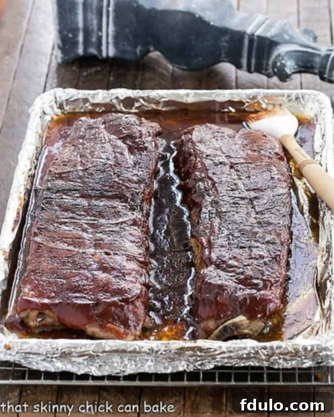 Two racks of baby back ribs on a sheet pan, generously coated with BBQ sauce, ready for broiling.