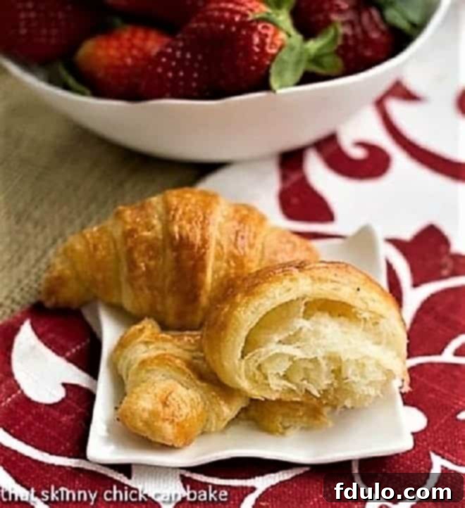 Freshly baked Classic Homemade Croissants arranged beautifully on a small white ceramic plate, accompanied by a vibrant bowl of ripe strawberries, highlighting their golden-brown perfection and inviting texture.