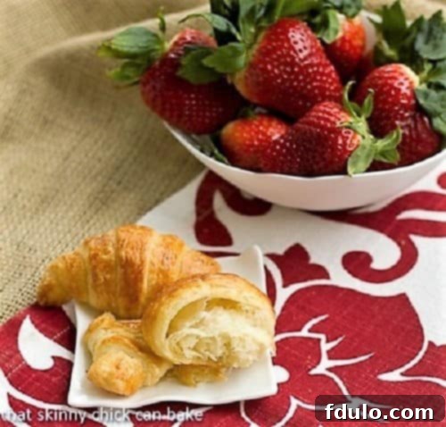 A close-up of a Homemade Classic Croissant, perfectly golden and flaky, with one croissant broken open to reveal its airy, layered interior. It's elegantly presented on a white plate alongside fresh strawberries, emphasizing its inviting texture and artisanal quality.