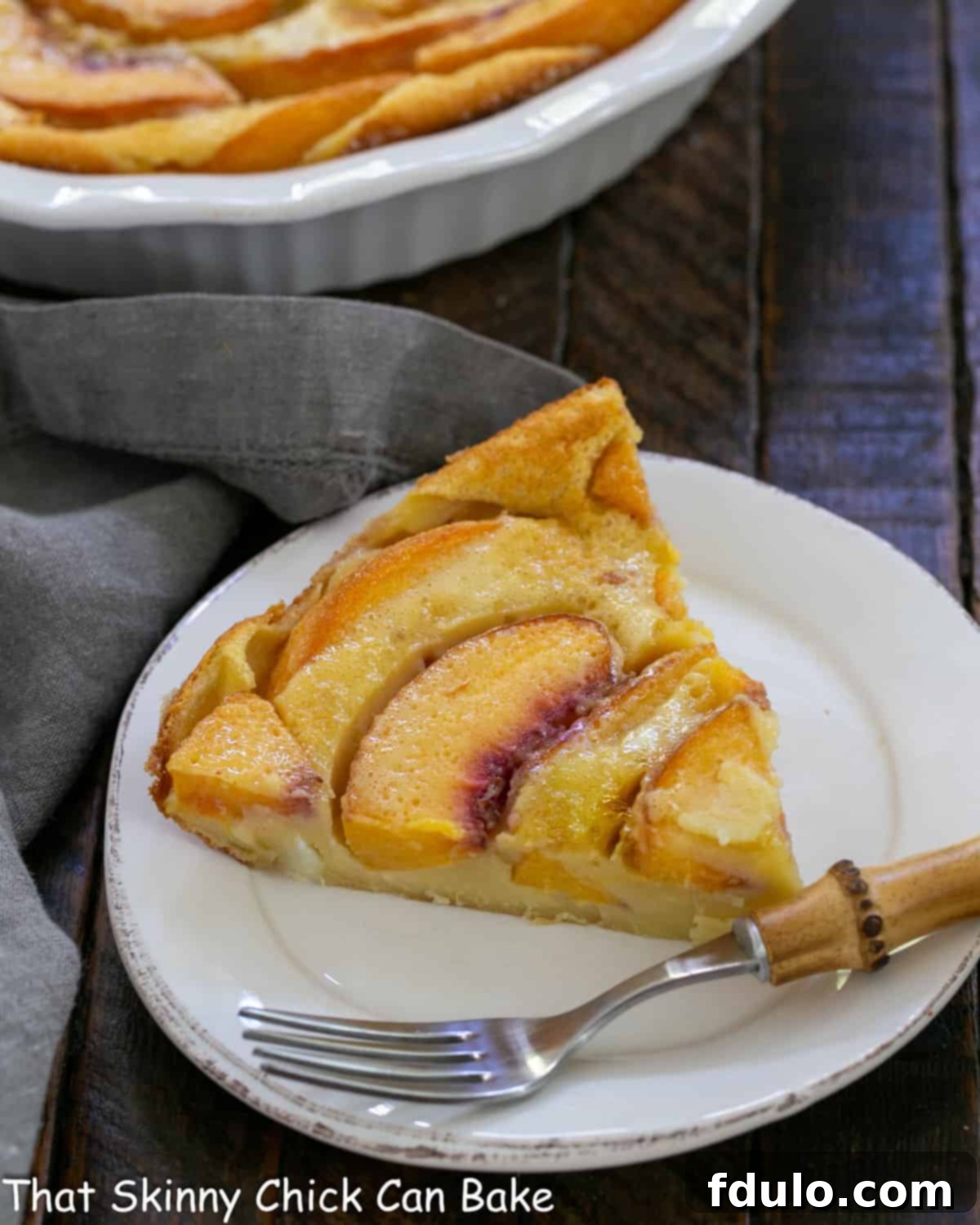 Fresh Peach Clafoutis - A classic French dessert with a peach twist! Slice of peach clafoutis on a white plate with a bamboo fork, showcasing its custardy texture and golden-brown top.