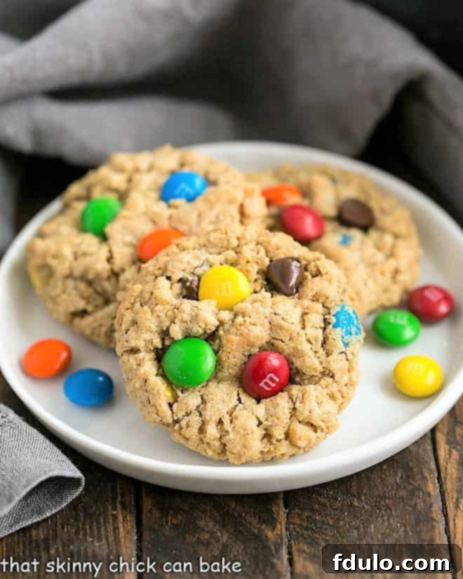 A stack of golden-brown Classic Monster Cookies on a white dessert plate, showcasing their chewy texture and colorful M&M's, chocolate chips, and walnuts.