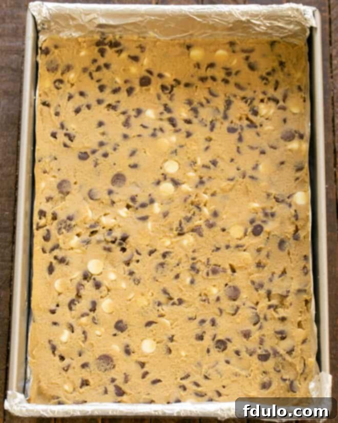 Chocolate chip cookie dough being spread evenly into a baking pan lined with foil or parchment paper.