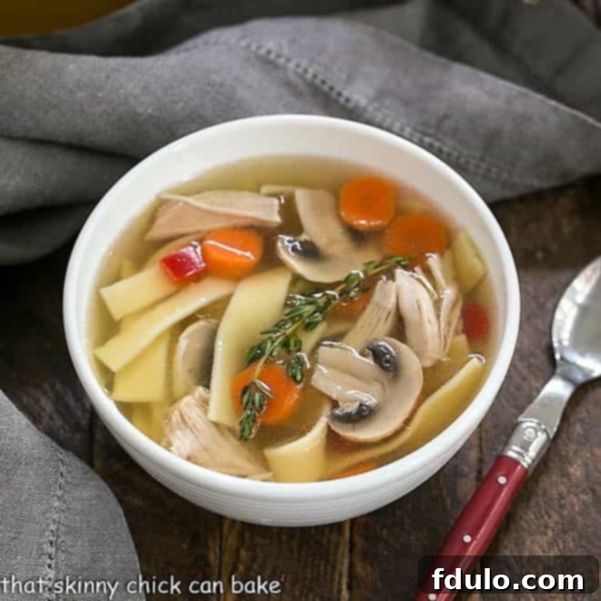 Overhead view of Homemade chicken stock, noodles and vegetables in a white bowl with a red handled spoon.