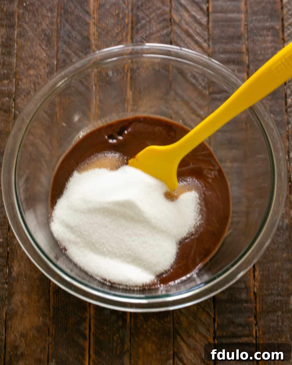 Step-by-step image showing granulated sugar and vanilla extract being stirred into the melted chocolate mixture.