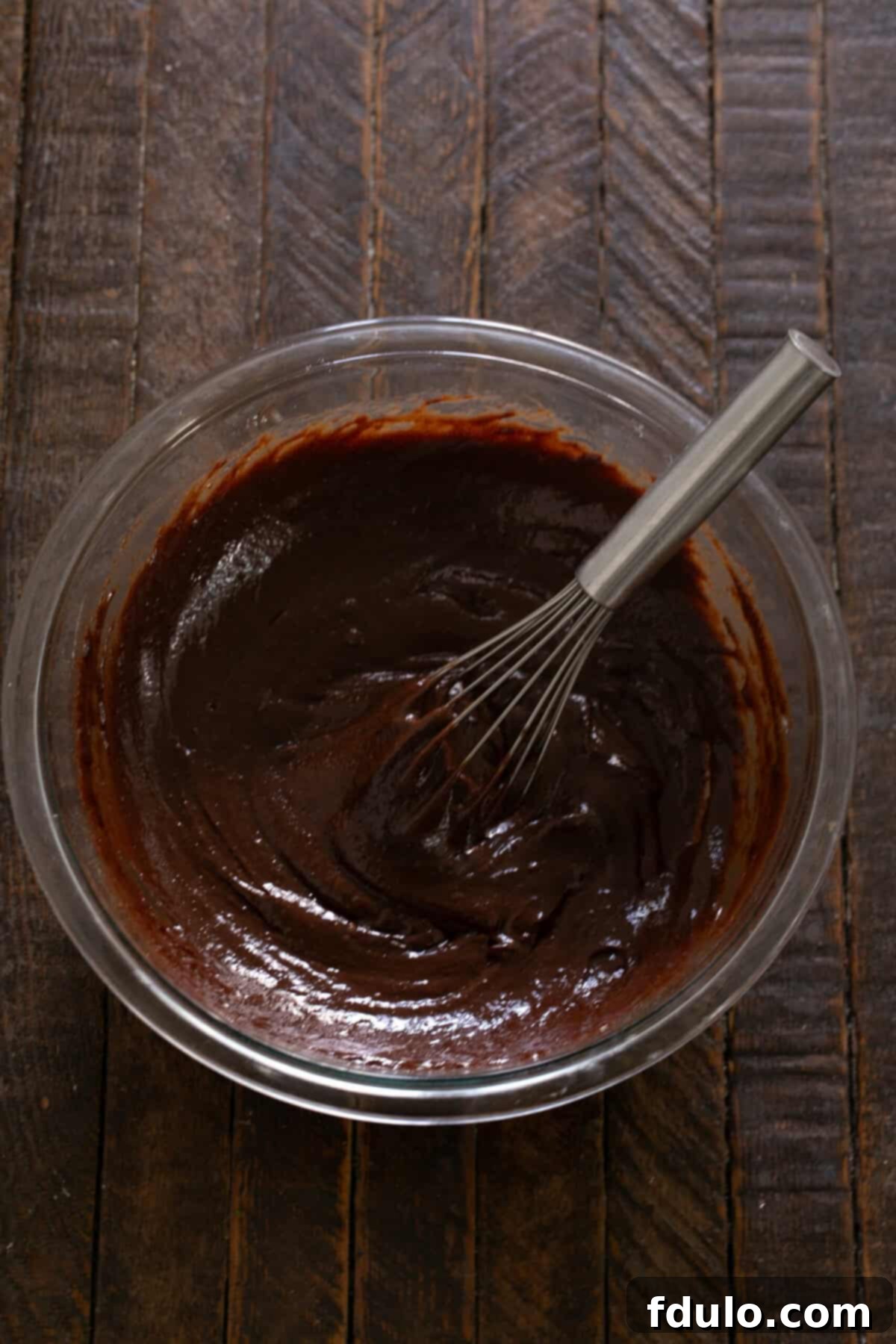 Step-by-step image illustrating the vigorous whisking of the brownie batter for one full minute until it becomes smooth and glossy.