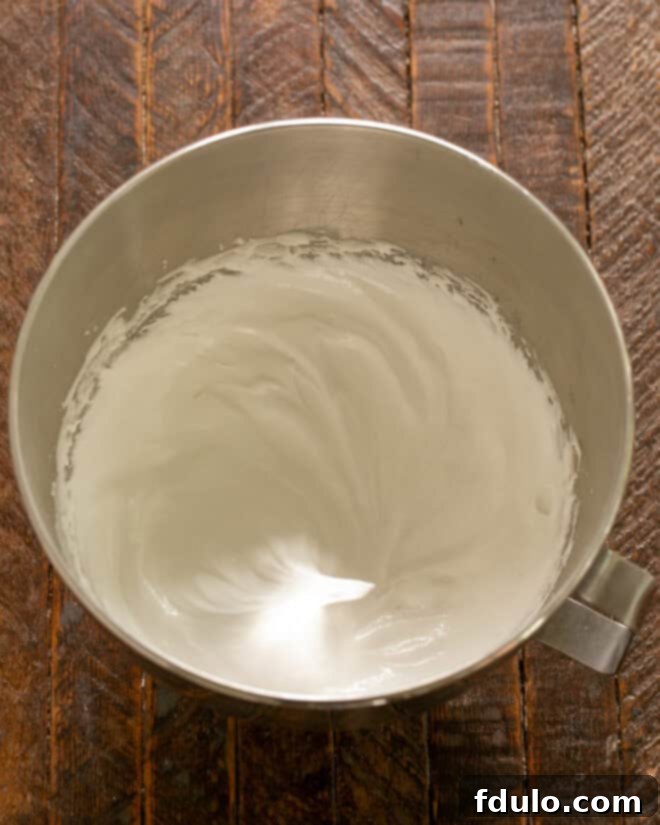 Superfine sugar being gradually added, one tablespoon at a time, to the beating egg whites in an electric mixer, creating a stiff and shiny meringue.