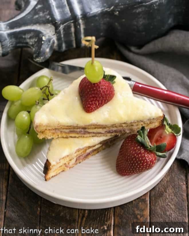 A beautifully presented Croque Monsieur Sandwich, sliced and stacked on a white plate, garnished with fresh grapes and strawberries, ready to be enjoyed.