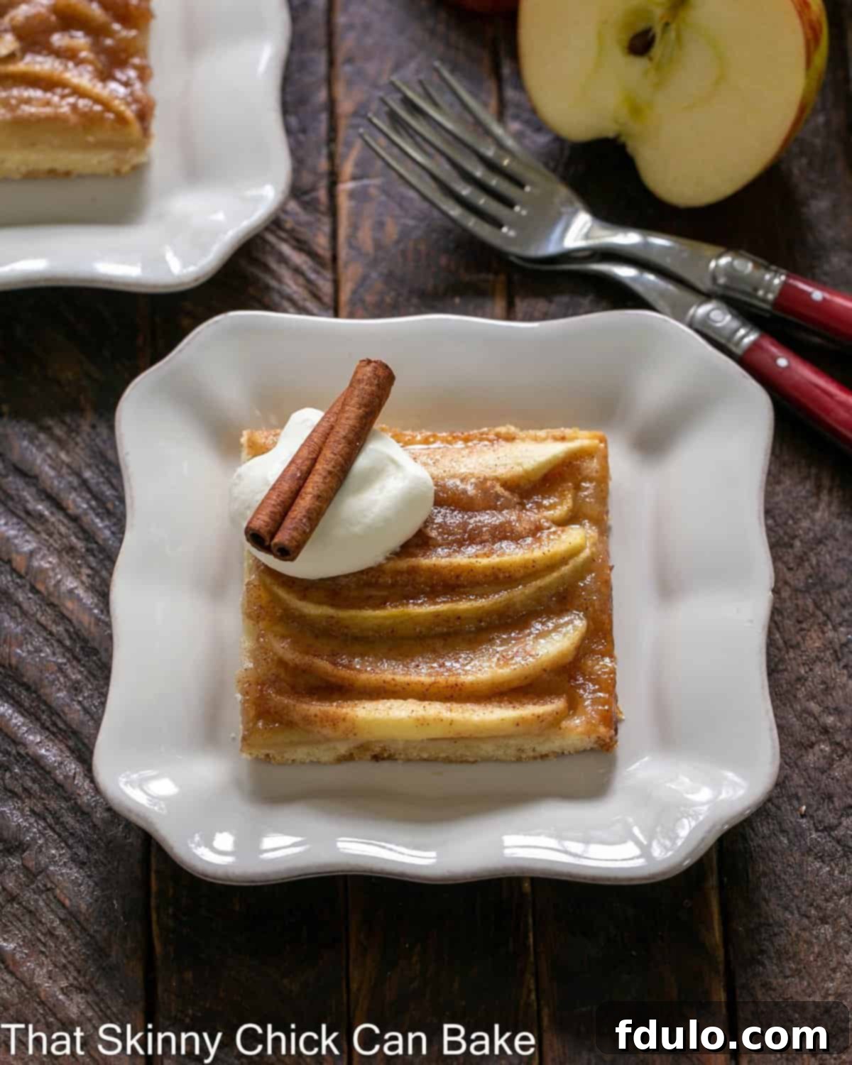A perfectly baked slice of German Apple Kuchen on a square white plate, garnished with a dollop of whipped cream and a cinnamon stick, ready to be enjoyed.