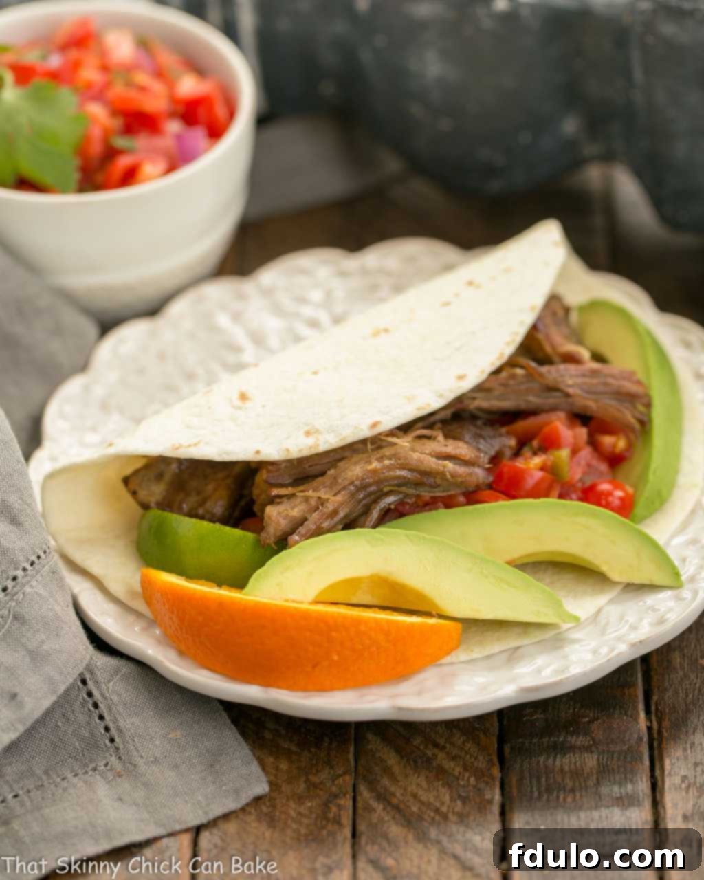 Slow cooked Pork Carnitas in a tortilla on a white plate with avocado and orange garnishes.