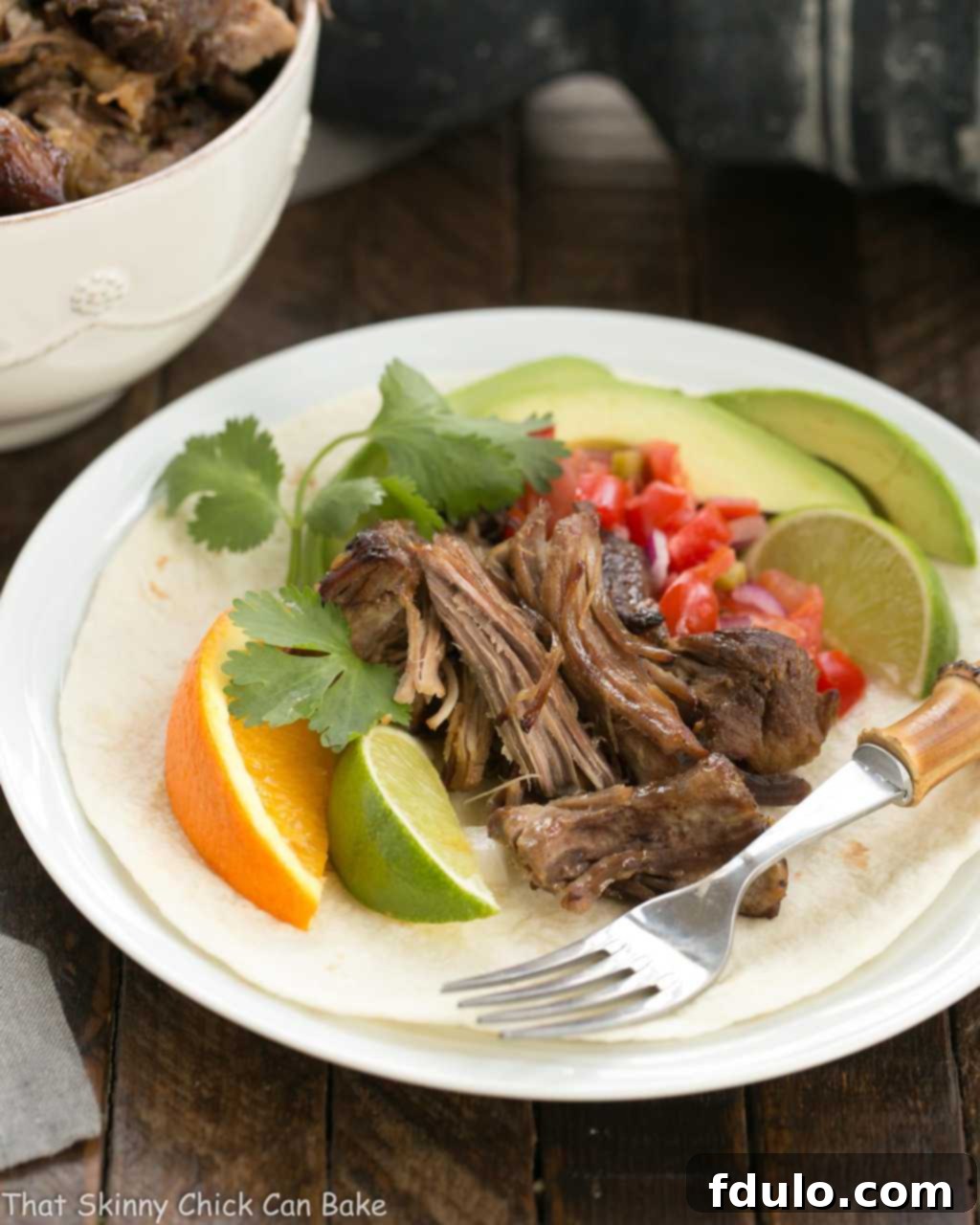 Slow cooked Pork Carnitas on a white plate with tomatoes, avocadoes and citrus.