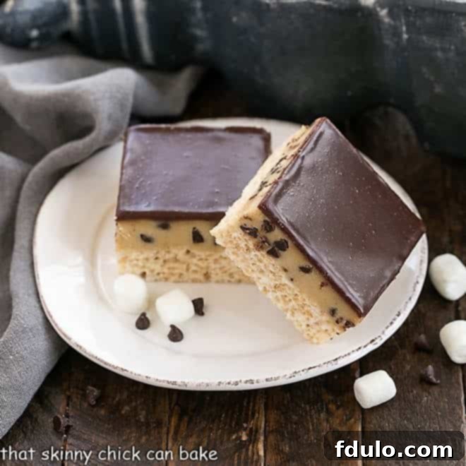 Two beautifully layered Cookie Dough Rice Krispie Treats, showing the golden crispy base, the light brown cookie dough, and the glossy dark chocolate ganache, on a white plate.