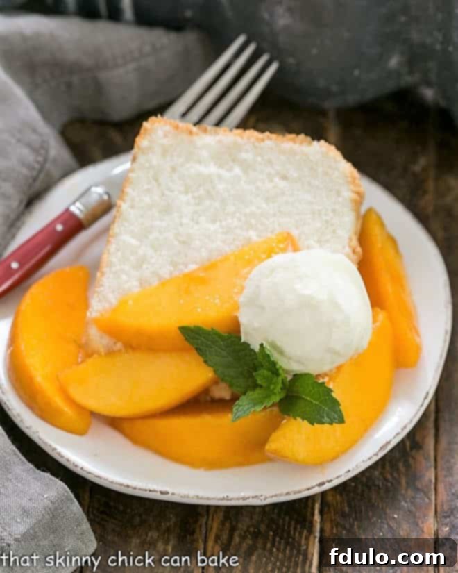 A slice of angel food cake on a dessert plate topped with fresh peach slices, whipped cream and mint.