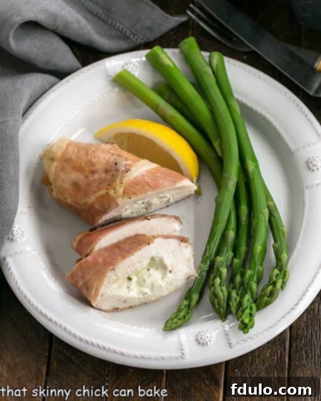 Baked Stuffed Chicken Breasts - Chicken breasts filled with creamy herbed Boursin cheese and wrapped in salty Prosciutto ham. An elegant entree that's easy to prepare! An overhead view of a beautifully presented Boursin Stuffed Chicken Breast, sliced to reveal its creamy filling, served alongside fresh lemon slices and vibrant green asparagus on a white dinner plate.