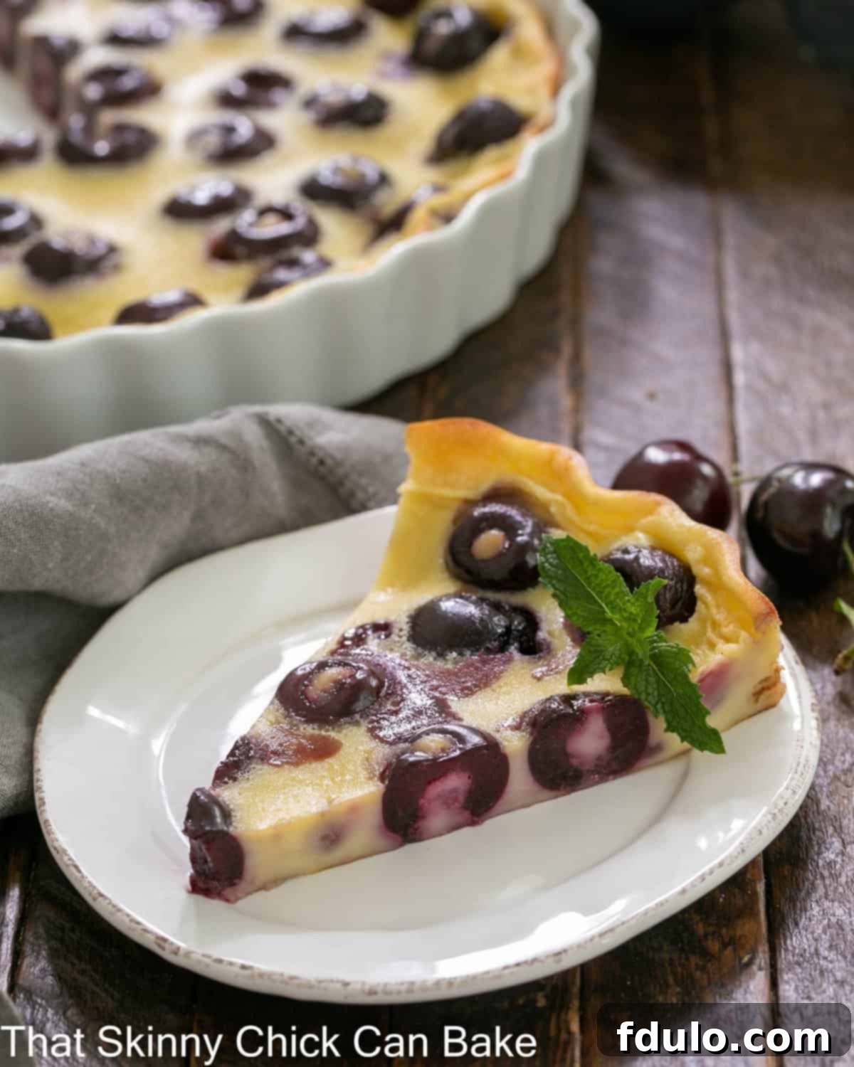 Slice of cherry clafoutis in front of the white ceramic baking dish.