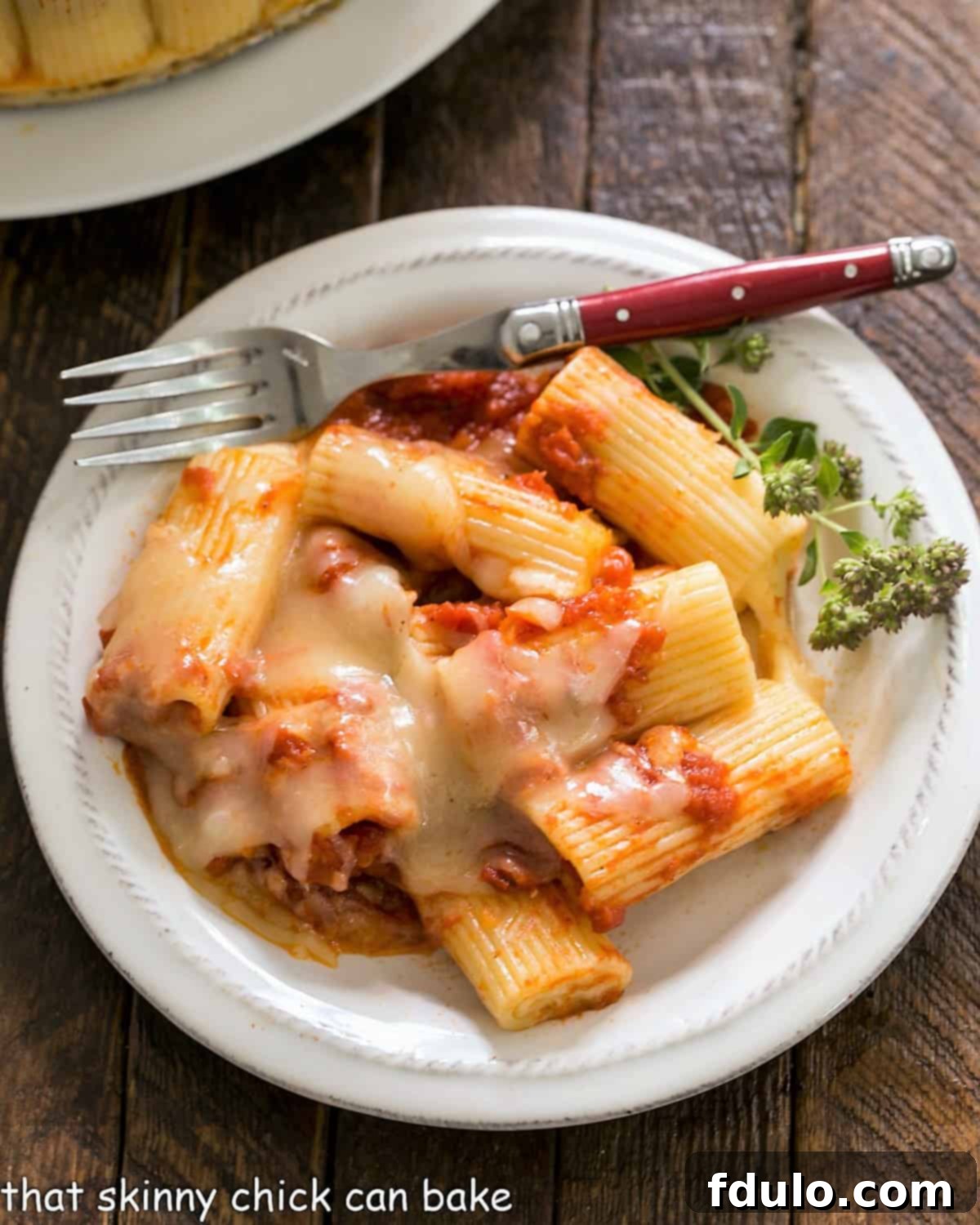 Overhead view of baked rigatoni on a small wite plate with a fork and herb garnish, highlighting the golden-brown cheese topping and vibrant sauce.