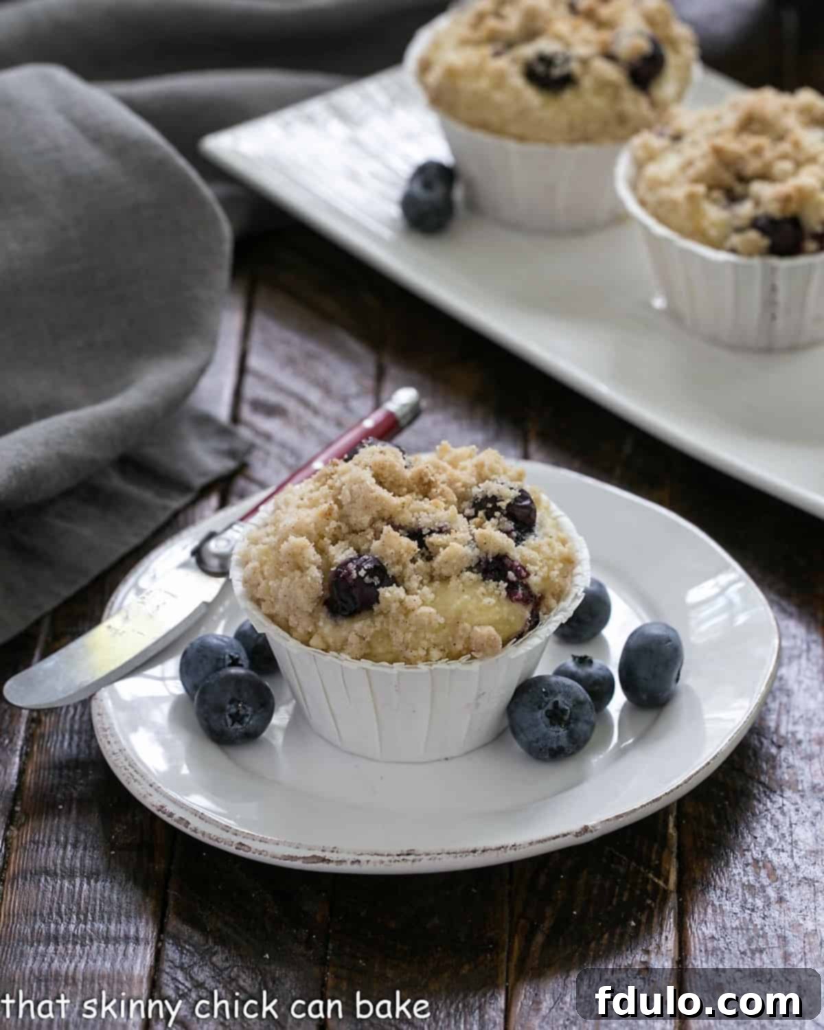 A single, perfectly baked Blueberry Cream Cheese Muffin sits on a white plate, accompanied by a small knife and a scattering of fresh blueberries, highlighting its delectable streusel crust.