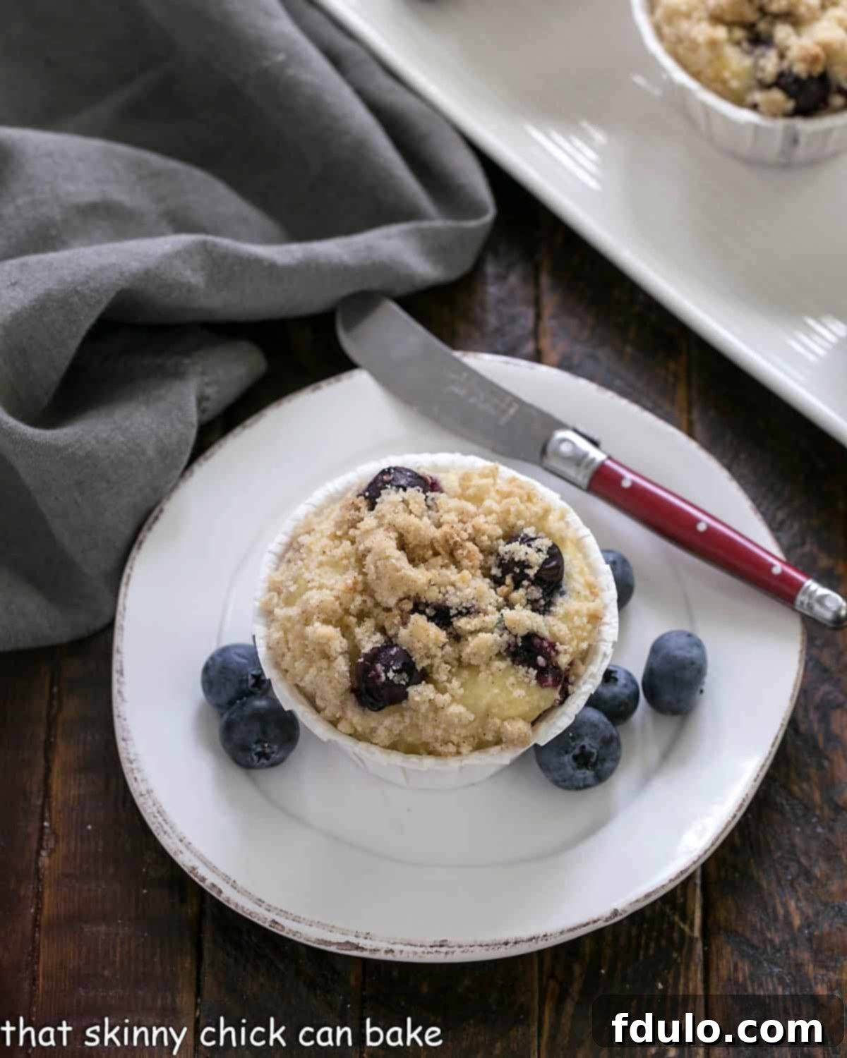 An overhead shot of a single Blueberry Cream Cheese Muffin on a white plate, paired with a knife featuring a rustic red handle, highlighting the muffin's appealing texture and streusel top.