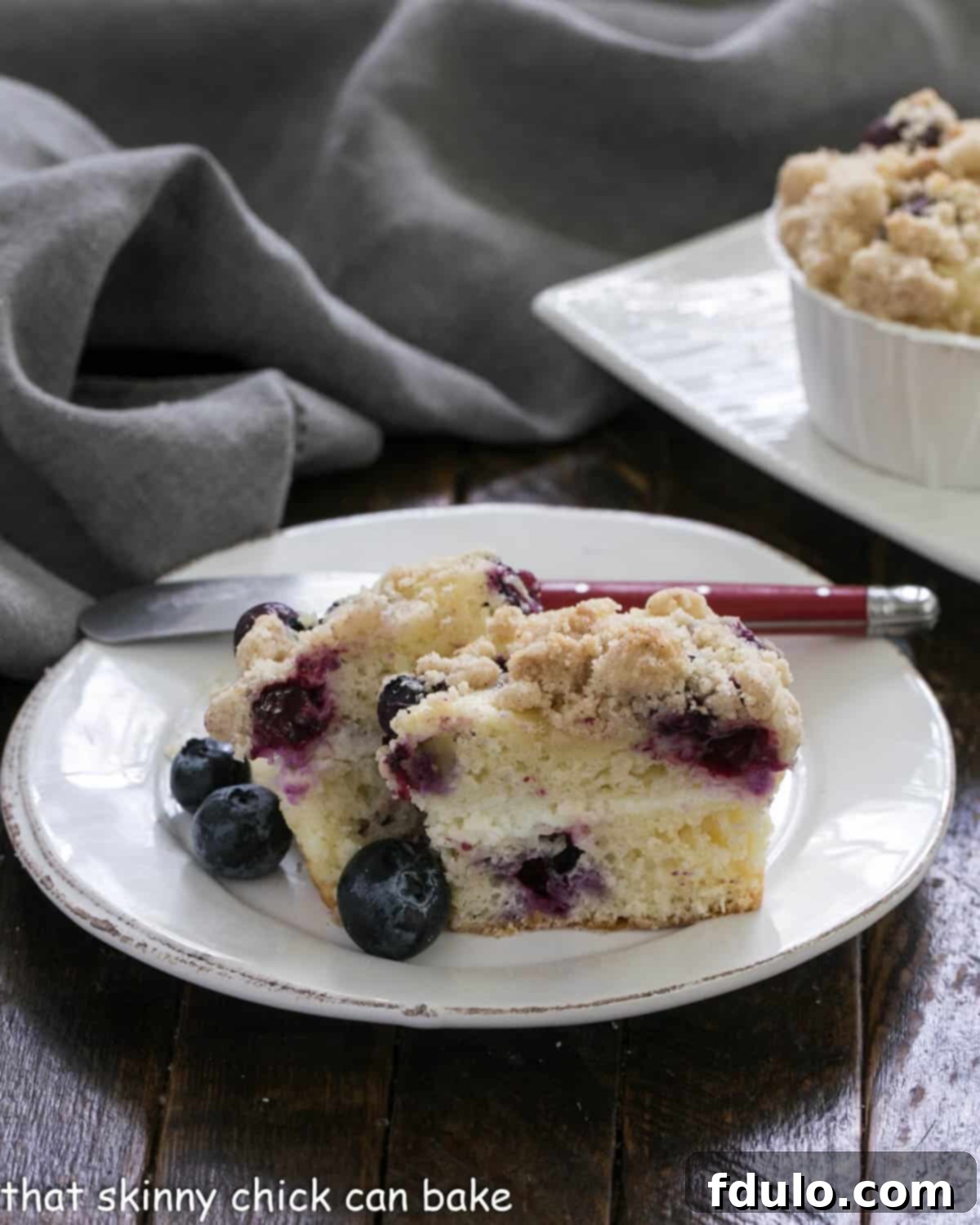 Two halves of a Blueberry Cream Cheese Streusel Muffin are displayed on a round white plate, revealing the luscious cream cheese filling and juicy blueberries within.