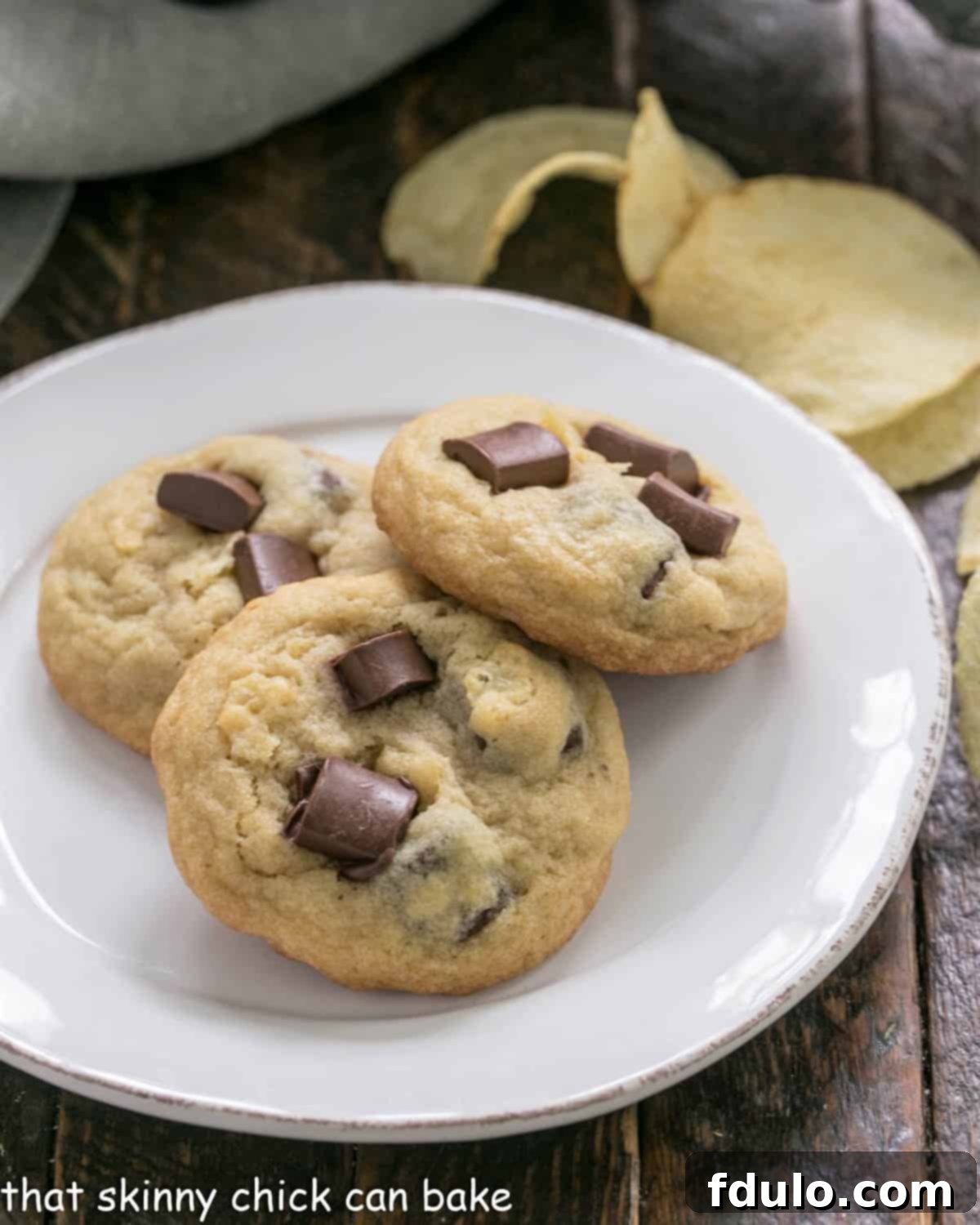 The Salty Sweet Chocolate Crunch Cookie 4 3 potato chip cookies on a white plate with potato chips in the background, emphasizing the key ingredient.