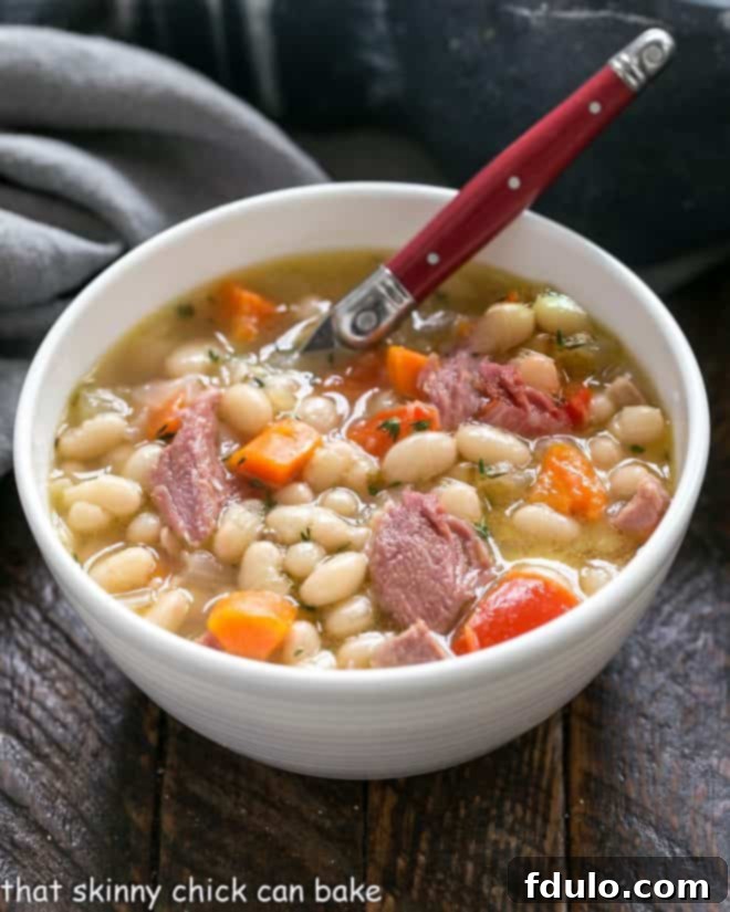 Hearty Navy Bean and Ham Soup 3 A close-up shot of old-fashioned Navy Bean Soup in a white bowl with a red-handled spoon, garnished with fresh herbs.