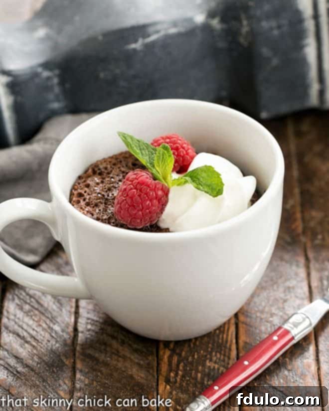 A beautifully presented chocolate mug cake in a white ceramic cup, adorned with a swirl of whipped cream, fresh berries, and a sprig of mint. A small spoon is gently placed beside the mug.