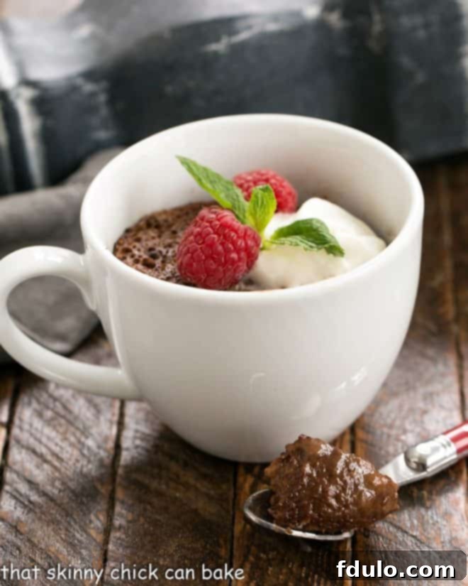 A close-up shot of a chocolate mug cake, beautifully garnished with whipped cream, fresh mint, and red berries. A spoon has just taken a scoop out of the cake, revealing its moist and tender interior.