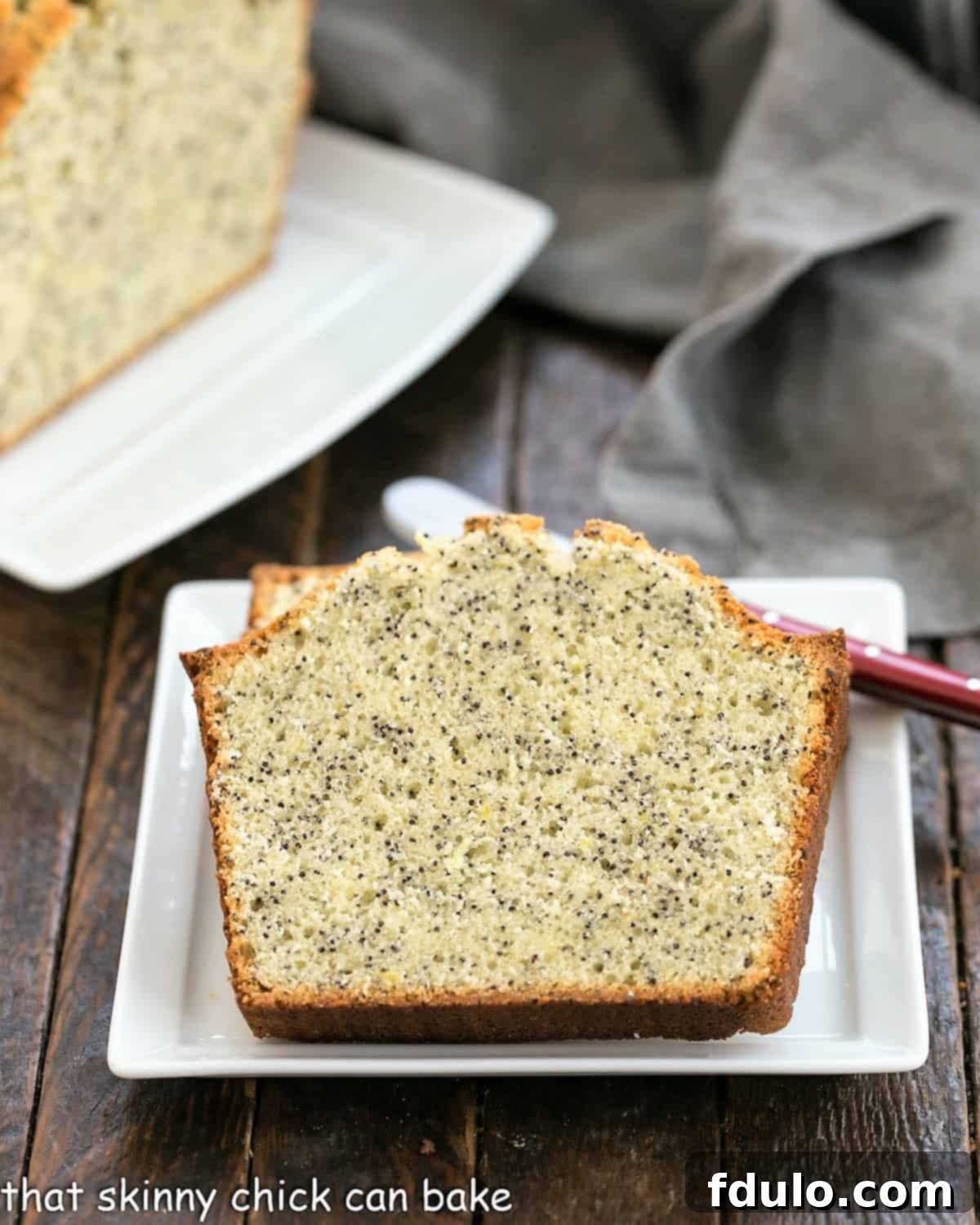 Two slices of poppy seed tea cake on a square white plate with a red handle knife in front of the rest of the loaf on a ceramic tray.